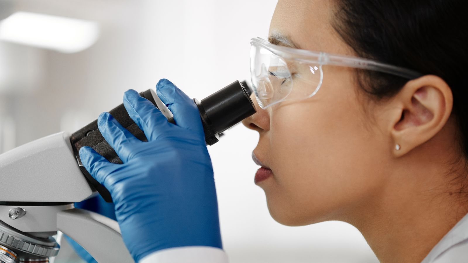 A scientist wearing safety glasses and gloves examines samples with a microscope in a laboratory setting.